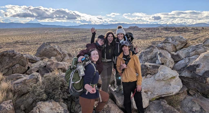 A group of people stand in front of large boulders and smile for the photo. Behind them, there is vast desert landscape, with mountains even further in the distance. 